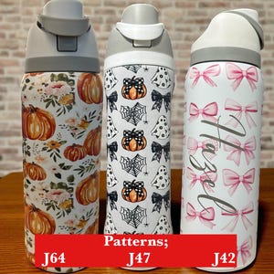 May include: Three water bottles with white and gray lids. One has a floral and pumpkin pattern, another has a Halloween theme with pumpkins and spiderwebs, and the third has pink bows and the name "Hazel".