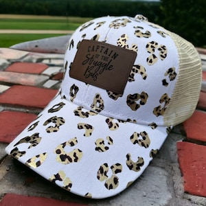 Op de afbeelding: Witte truckercap met een gouden luipaardprint en een bruin leren patch met de tekst "Captain of the Struggle Bus".