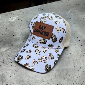 Op de afbeelding: Witte baseballpet met een luipaardprint en gouden accenten. De pet heeft een bruin leren patch met de tekst "DAY DRINKER" erop.