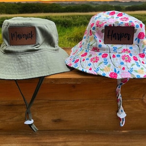 May include: Two children's bucket hats. One is green with the name "Maverick" on it. The other is white with a floral pattern and the name "Harper" on it.