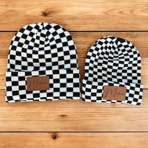 Pode incluir: Dois gorros de malha xadrez preto e branco com patches de couro marrom. O gorro maior tem a palavra "Mama" no patch e o gorro menor tem a palavra "Mini" no patch.