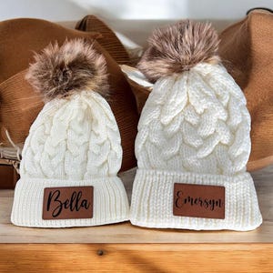 May include: Two white knit hats with faux fur pom poms and brown leather patches. The hats have the names "Bella" and "Emersyn" written on the patches.