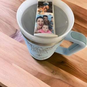 May include: A light blue travel mug with a white lid and a white photo strip insert. The photo strip features a family of four. The mug has a white and silver design on the side.