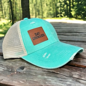 Può includere: Un cappello da camionista in rete turchese e beige con una toppa in pelle marrone che dice "DAY DRINKER" in lettere nere.