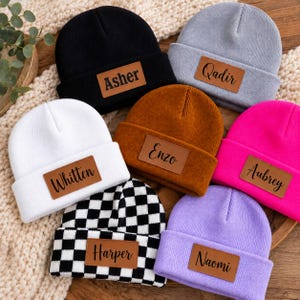 Puede incluir: Una colección de siete gorros de punto en negro, gris, blanco, marrón, rosa intenso, lavanda y un patrón de cuadros blanco y negro. Cada gorro tiene un parche rectangular de piel sintética con un nombre impreso en letra cursiva.