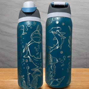 Op de afbeelding: Twee turquoise waterflessen met zilveren illustraties van haaien, vissen en bellen. De flessen hebben een grijs en wit deksel met een drukknopopening. De flessen staan op een houten oppervlak.