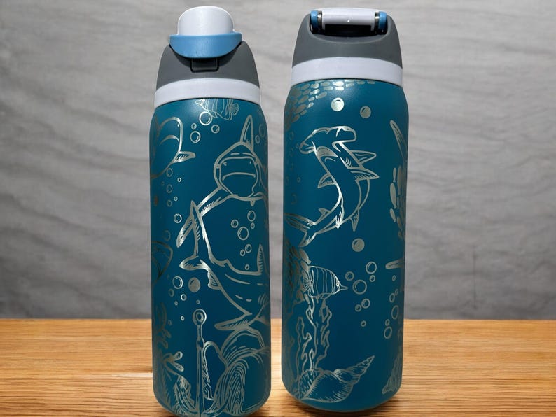 Op de afbeelding: Twee turquoise waterflessen met zilveren illustraties van haaien, vissen en bellen. De flessen hebben een grijs en wit deksel met een drukknopopening. De flessen staan op een houten oppervlak.