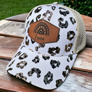 Può includere: Un cappello da camionista con stampa leopardata bianca e marrone con una toppa in pelle marrone che presenta un arcobaleno e il nome "Luna".