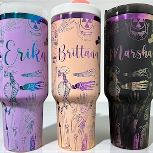 Könnte beinhalten: Drei personalisierte Becher mit einem Skelett-Thema. Die Becher sind in den Farbtönen Lila, Pfirsich und Schwarz gehalten. Jeder Becher hat einen Namen in Schreibschrift und ein sich wiederholendes Muster aus Skelettelementen. Die Deckel sind weiß mit einem metallischen Rand.