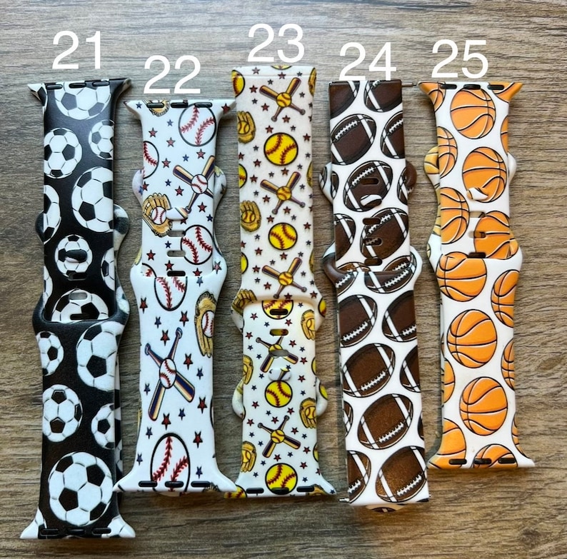 May include: Five different Apple Watch bands with various sports-themed patterns. The bands are white with black, brown, or orange accents. The patterns include soccer balls, baseballs, softballs, footballs, and basketballs.