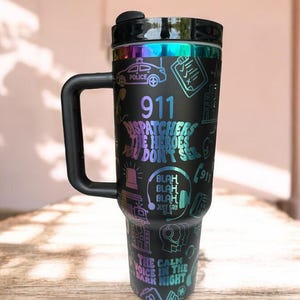 Può includere: Tazza da viaggio nera con finitura arcobaleno iridescente. La tazza presenta un design con un'auto della polizia, una sirena, un auricolare e il testo "911 Dispatchers The Heroes You Don't See".