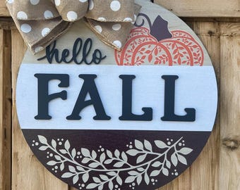 May include: A round wooden sign with a brown and white polka dot bow. The sign has the words "hello FALL" in black, a pumpkin design, and a decorative leaf border. The sign is on a wooden background.