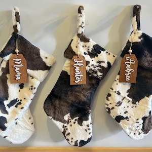 May include: Three cow print Christmas stockings with wooden tags that say "Mama", "Hunter", and "Aubree".