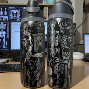 May include: Two black water bottles with gray lids, adorned with a silver skeletal design. The bottles feature a variety of human bone illustrations, including skulls, ribs, and limbs. The bottles are designed for hydration.