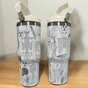 May include: Two white stainless steel travel tumblers with a black and white anatomical design. The tumblers have a white lid with a black handle.