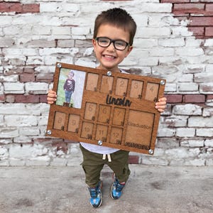 May include: A rectangular wooden school year photo frame with twelve numbered slots for grades kindergarten through twelfth grade, featuring the name Lincoln in script, a graduation photo space, and metal corner fasteners for assembly.