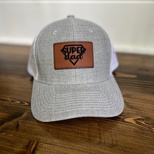 May include: A gray and white trucker hat with a brown leather patch that says "super dad" in black lettering.