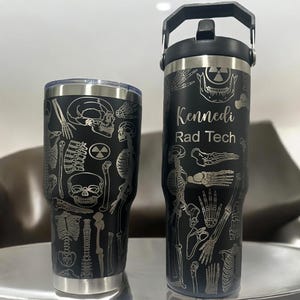 Pode incluir: Dois copos de aço inoxidável preto com um design esquelético e um símbolo de radiação. O copo maior tem uma alça e o texto "Kennedi Rad Tech" gravado no lado.