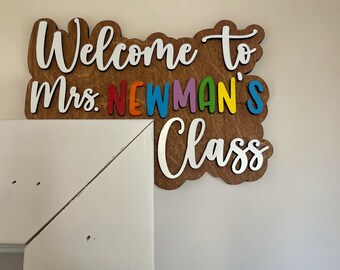 Może przedstawiać: Tabliczka powitalna z napisem "Welcome to Mrs. Newman's Class" w białym kolorze na brązowym drewnianym tle. Litery w "Newman's" są kolorowe.