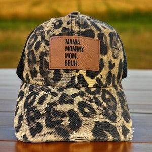 Può includere: Un cappello da camionista con stampa leopardata nero e marrone con una toppa in pelle marrone che dice "MAMA. MOMMY. MOM. BRUH."