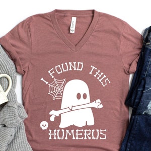 May include: A mauve V-neck t-shirt with a white graphic of a ghost holding a bone with the text "I found this humerus".