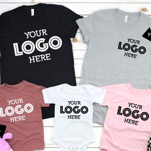May include: A collection of t-shirts and a onesie in various colors, including black, gray, red, white, and pink. Each garment features the text "YOUR LOGO HERE" in a bold, black font. The shirts have different print styles.