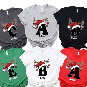 May include: Six Christmas themed t-shirts with different letters and names. The shirts are in various colors, including grey, black, green, white, and red. Each shirt features a reindeer with a Santa hat and ornaments.