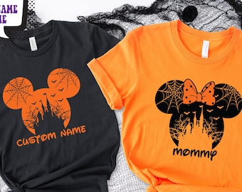May include: A black t-shirt with an orange Mickey Mouse silhouette featuring a spiderweb, bats, and castle graphic with the text CUSTOM NAME, and an orange t-shirt with a black Mickey Mouse silhouette featuring a bow, spiderweb, bats, and castle graphic with the text mommy.