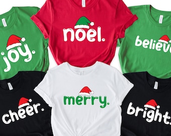 May include: A collection of five short-sleeved t-shirts in red, white, green, and black. Each shirt features a holiday-themed word printed in white or green, including joy, noel, believe, cheer, merry, and bright, topped with a small Santa hat.