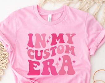 Könnte beinhalten: Pinkes T-Shirt mit dem Text "IN MY CUSTOM ERA" in einer Retro-Schriftart mit pinkfarbenen und weißen Glitzerakzenten.