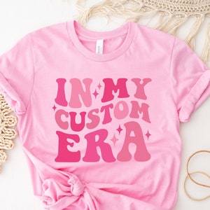 May include: Pink t-shirt with the text "IN MY CUSTOM ERA" in a retro font with pink and white glitter accents.