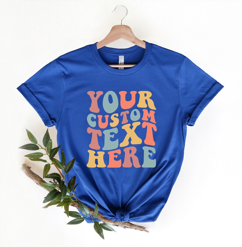 May include: Royal blue t-shirt with rolled-up sleeves, hanging on a wooden hanger. The shirt features the text "YOUR CUSTOM TEXT HERE" in a retro, colorful font. A sprig of greenery rests below the shirt.
