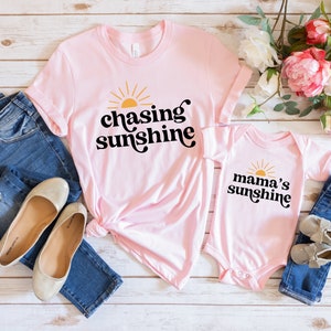 May include: Pink t-shirt with the text "chasing sunshine" in black with a yellow sun graphic. A pink bodysuit with the text "mama's sunshine" in black with a yellow sun graphic.