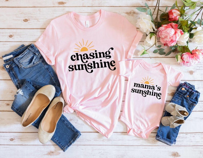 May include: Pink t-shirt with the text "chasing sunshine" in black with a yellow sun graphic. A pink bodysuit with the text "mama's sunshine" in black with a yellow sun graphic.