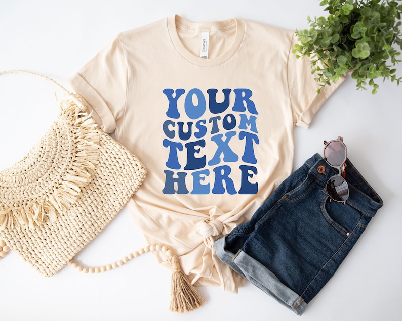 May include: A cream-colored t-shirt with the text "YOUR CUSTOM TEXT HERE" in shades of blue. The shirt is styled with denim shorts, sunglasses, and a woven bag, suggesting a casual, everyday look.