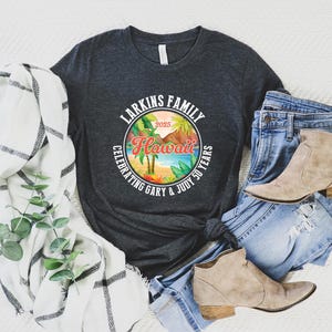 May include: A dark gray t-shirt with a colorful Hawaii graphic, jeans, and tan ankle boots. The graphic reads "Larkins Family Hawaii Celebrating Gary & Judy 50 Years 2025". A white and black plaid scarf and greenery are also in the image.