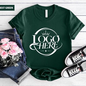 May include: Forest green t-shirt with a white logo that says "LOGO HERE" in a circular design. The shirt is paired with blue jeans, pink roses, and black and white sneakers.