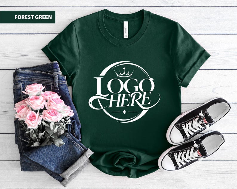 May include: Forest green t-shirt with a white logo that says "LOGO HERE" in a circular design. The shirt is paired with blue jeans, pink roses, and black and white sneakers.