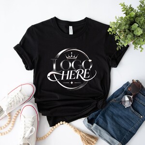 May include: Black t-shirt with a white and silver logo that says "LOGO HERE" in a circular design with a crown. The shirt is paired with white sneakers, denim shorts, and sunglasses. A sprig of greenery and a beaded necklace are also in the image.