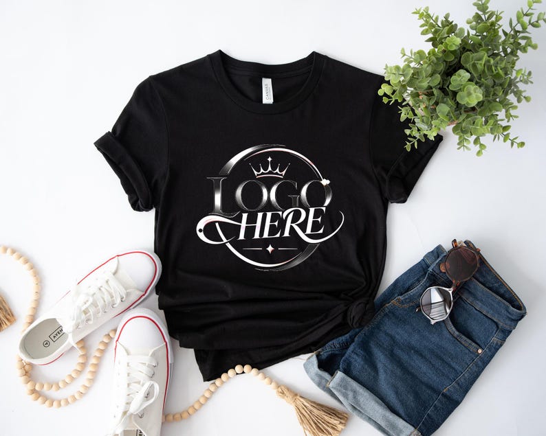 May include: Black t-shirt with a white and silver logo that says "LOGO HERE" in a circular design with a crown. The shirt is paired with white sneakers, denim shorts, and sunglasses. A sprig of greenery and a beaded necklace are also in the image.