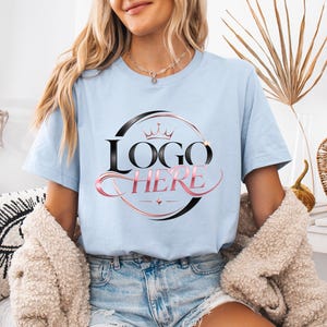 May include: Light blue t-shirt with a black and pink logo that says "LOGO HERE". The logo is inside a black circle with a crown on top. The model is wearing denim shorts and a beige jacket.