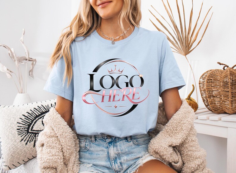 May include: Light blue t-shirt with a black and pink logo that says "LOGO HERE". The logo is inside a black circle with a crown on top. The model is wearing denim shorts and a beige jacket.