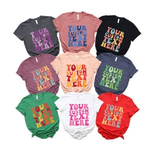 May include: A collection of nine t-shirts in various colors, each featuring the text "YOUR CUSTOM TEXT HERE" in a retro, wavy font. The shirts are in shades of gray, pink, black, burgundy, peach, navy, green, white, and red.