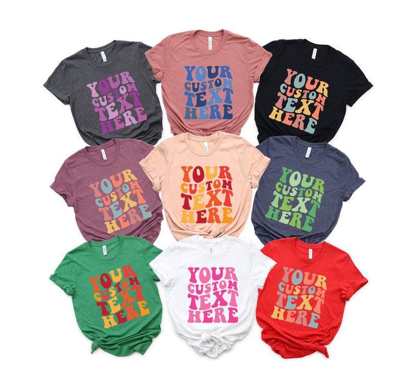 May include: A collection of nine t-shirts in various colors, each featuring the text "YOUR CUSTOM TEXT HERE" in a retro, wavy font. The shirts are in shades of gray, pink, black, burgundy, peach, navy, green, white, and red.