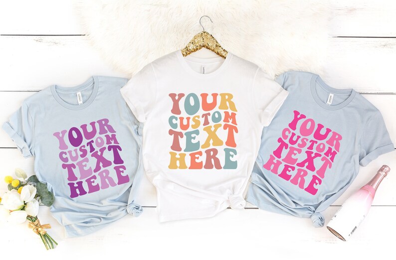 May include: Three t-shirts with the text "YOUR CUSTOM TEXT HERE" in a retro font. The shirts are light blue and white. The text is in various colors, including pink, purple, orange, and teal. The shirts are displayed on a white surface.