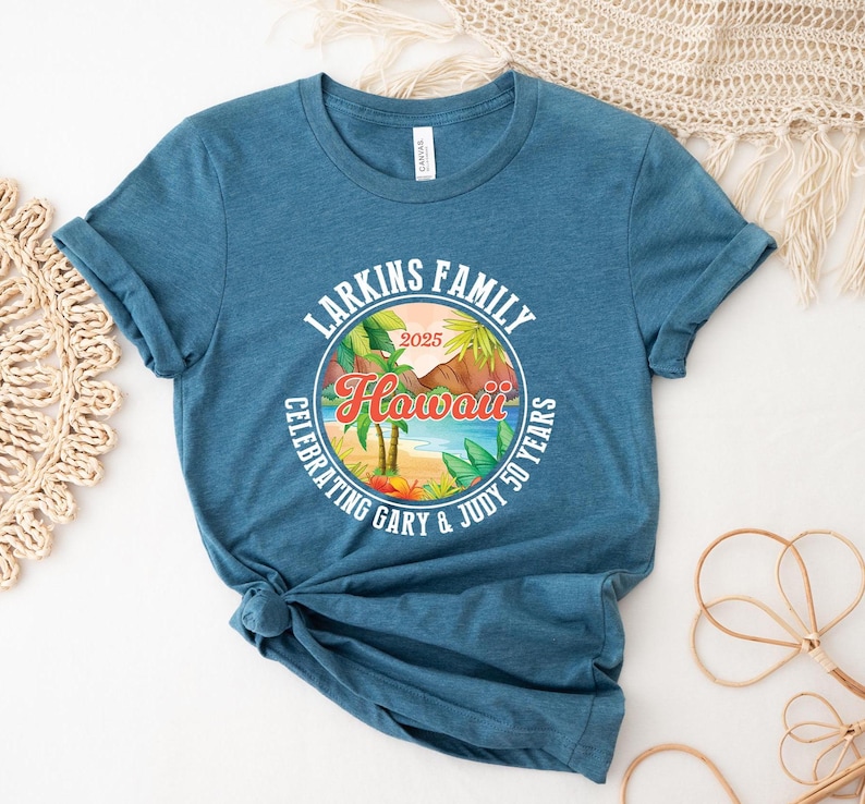 May include: Teal-colored t-shirt featuring a circular graphic with the text "Larkins Family Hawaii 2025 Celebrating Gary & Judy 50 Years." The graphic depicts a tropical beach scene with palm trees, mountains, and ocean. The shirt has rolled-up sleeves.