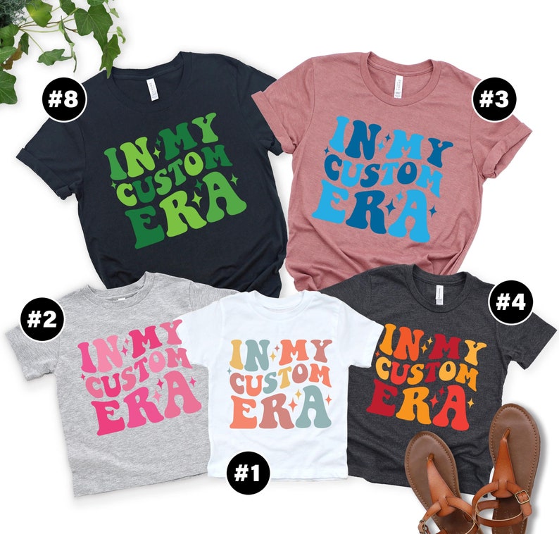 May include: Five t-shirts with the text "In My Custom Era" in different colors and fonts. The shirts are arranged in a row, with the largest shirt in the middle and the smallest shirts on either end.