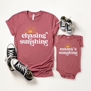 May include: Matching shirts for a parent and child. The adult shirt is maroon and says "chasing sunshine" with a yellow sun graphic. The baby bodysuit is maroon and says "mama's sunshine" with a yellow sun graphic.