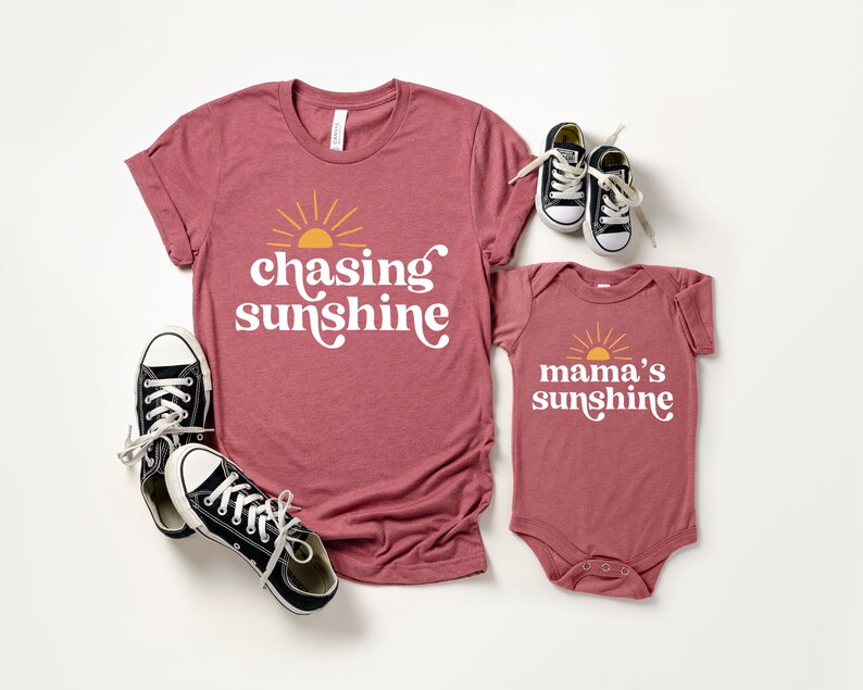 May include: Matching shirts for a parent and child. The adult shirt is maroon and says "chasing sunshine" with a yellow sun graphic. The baby bodysuit is maroon and says "mama's sunshine" with a yellow sun graphic.