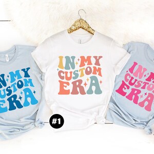 May include: Three t-shirts with the text "In My Custom Era" in a retro font with stars and different color schemes. The shirts are light blue, white, and pink.